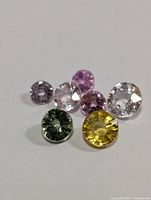 Six small round cut fancy color sapphire gemstones arranged on a white surface, showing various colors including yellow, green, purple, and clear.