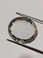 Oval shaped green amethyst gemstone held by tweezers, showing faceted top view, translucent light green color.