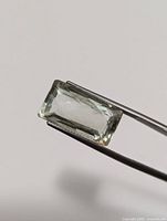 Close-up photo of the rectangular cut light green transparent gemstone held by tweezers, showing its color and clarity.