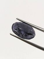 Close-up image of single oval faceted blue-violet Iolite gemstone held in tweezers showing surface details and inclusions.
