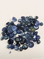 Collection of loose genuine sapphire gemstones arranged on a white background showing variety of sizes, shapes, and blue color shades.