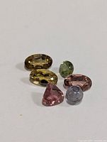 Six faceted tourmaline gemstones of different colors arranged on a white background, showing variety in shape and color.
