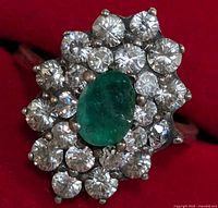 Close-up front and angled view of silver ring with oval emerald surrounded by cubic zirconia stones on a red background.