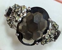 Close-up of the front and side of a silver ring featuring a large oval faceted smokey quartz stone held by prongs, flanked by clusters of small green stones on silver band detailing.