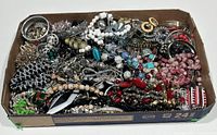 Top view showing the full assortment of costume jewellery items in an open cardboard box, including bangles, beaded necklaces, and various pendants and rings, colorfully mixed and layered.