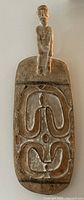 Front view of wooden tray or wall hanging showing carved figure on handle with abstract curvilinear patterns on the main surface.
