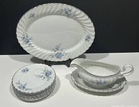 Photo displaying all pieces in the set: 7 bread and butter plates stacked, oval serving platter behind, and gravy boat with underplate to right. All pieces show the blue floral pattern with silver trim on white ironstone.