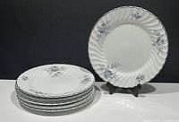 Seven white Johnson Bros Ironstone Snowhite Regency Blue floral dinner plates stacked with one plate displayed upright on a stand, showing scalloped edges and floral designs.