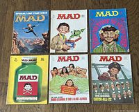 Top-down view of six Mad magazines from the 1960s-1970s, arranged in two rows of three on a wood floor, showing different detailed colorful covers.