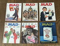 Six MAD magazines laid out in two rows on a wooden surface, showing full covers and different themes of satire and parody.