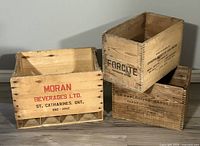 Three wooden crates: one light wood Moran Beverages Ltd. 12 bottle crate on the left with red and black text, and two darker Forcite wooden crates stacked on the right with black printed warnings.