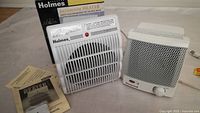 Two indoor heaters shown together on a surface with manuals and box visible, one is Holmes bathroom heater and the other is smaller compact heater.