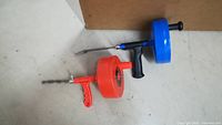 Two manual drain snakes, one red and one blue, placed on a floor with a cardboard piece against the wall.