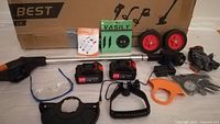 Battery electric weed wacker with attachments and accessories laid out including batteries, charger, goggles, wheels, and replacement parts with boxed packaging in background.