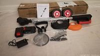 Lot laid out on floor showing all parts and accessories for battery weed wacker: main unit, battery, charger, wheels, blades, handle, guards and brackets.