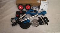 Photo showing battery powered weed wacker disassembled parts including batteries, charger, saw blade, wheels, and manual.