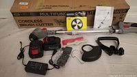 Battery powered cordless brush cutter and weed wacker with battery, charger, blade, manual, and attachments laid out in front of original packaging box