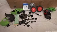 Top-view photo showing all components laid out: weed wacker body, battery, charger, wheels, manual, green guard, several metal cutting blades, and attachment parts.