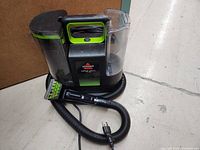 Photo of the Bissell Little Green Max Pet Carpet Cleaner showing the main unit, hose, power cord, and cleaning tool attachment on the floor against a wall.