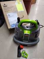 Photo of Bissell Little Green ProHeat carpet cleaner with hose and cleaning attachment in front of original box and instructions.
