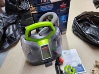Bissell Little Green ProHeat portable carpet cleaner unit with green handle and transparent tanks, connected to grey flexible hose with red-bristle brush, placed on floor beside original product box and instruction manual.