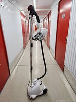 White Conair upright garment steamer with accessories, placed in a hallway. The steamer has a wheeled base, metal telescoping pole, hose to steam nozzle, and accessory attachments wrapped in plastic.
