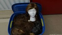 Plastic bin containing multiple wigs of different colors and styles along with a white styrofoam mannequin head for wig display.