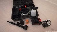 Electric cordless high pressure cleaning machine components displayed outside of molded carrying case on light surface. Items include spray gun, hose, foam bottle, battery, charger, and multiple nozzles.