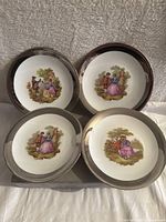 Photo showing four 7-inch porcelain plates with silver trim arranged on fabric background, each plate featuring detailed romantic couple scenes in pastel colors.