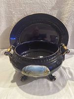 Blue porcelain tureen with two handles, three decorative feet, and oval painted landscape scene, in front of matching blue platter with gold trim