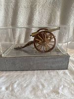 Front side view of brass and wood model cannon inside clear display case on wooden base.