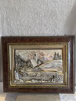 Front view of framed metal and wood art showing village scene with church, trees, mountains, and stone bridge.