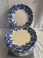 Stack of 11 white porcelain plates with blue fruit and floral decorative border, showing pattern and scalloped edges.