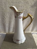 Front side view of the white porcelain pincher vase with gold handle and floral details against a grey fabric background.