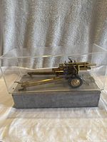 Side angle of the gold finished metal model cannon inside clear display case on gray base, showing wheels, barrel, and support legs.