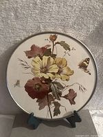 Full view of the round porcelain tray on a stand, showing floral artwork with a butterfly and bee, bordered by metal rim.