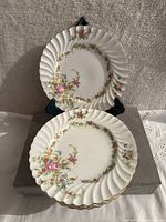 Two porcelain plates, front view showing scalloped edges with gold trim and colorful floral design around the inner rim.