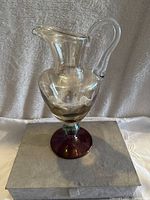 Full view of clear glass vintage pitcher with a deep red base, showing curved handle and spout, placed on grey cloth surface.
