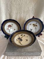 Three vintage porcelain plates with floral and bird designs displayed upright and flat, showing detailed gold accents on dark blue borders.
