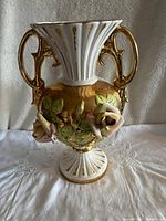 Front view showing two gold handles, porcelain rose decorations, and hand-painted green leaves on gold background with fluted neck and base.