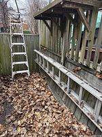 View showing both ladders: 7-foot step ladder standing upright and 10-foot extension ladder resting horizontally against wooden railing covered with leaves.