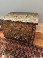 Angled side view showing embossed relief scene on the side of the brass box, with human figures and decorative border.