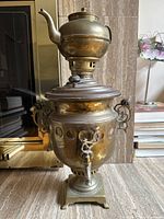 Large vintage brass tea urn standing upright on a stone floor near a fireplace.