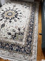 Full view of rectangular carpet showing central floral medallion pattern and multi-layered borders in navy, beige, olive, and cream colors positioned on hardwood floor.
