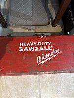 Closed red metal carrying case with white text reading 'Heavy Duty Sawzall Milwaukee' showing wear and scratches.