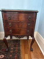Front view of one dark wood side table with glass top and three drawers. Shows metal ring drawer pulls and ball claw feet.