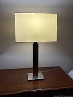 Full view of the metal table lamp turned on, showing rectangular beige fabric shade and polished metal base and shaft.