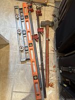 Overview of the tools showing two spirit levels (one metal and silver, one orange plastic), two black metal bar clamps with red handles, and a sledgehammer with a wooden handle and large metal head, all laid on tiled floor.
