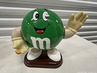 Front view of Ms. Green M&M candy dispenser showing face, raised waving hand, orange button, base