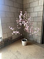 Artificial cherry blossom plant with multiple pink flowered branches in a white pot, placed against a concrete block wall.